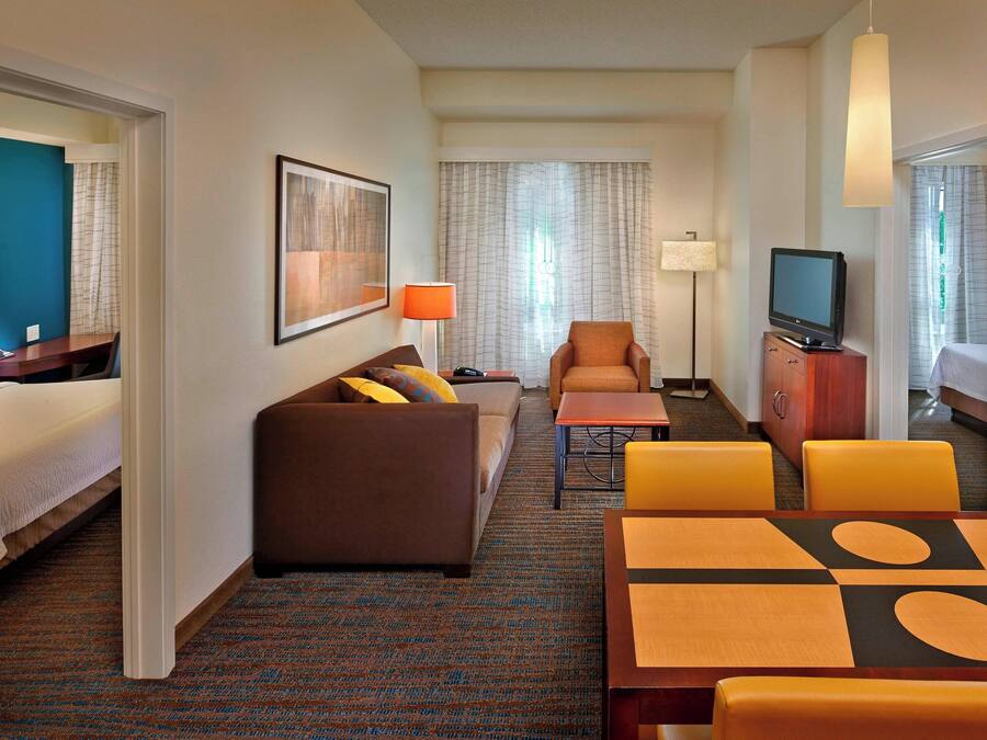 A hotel room with a bed, a sofa, a dining table with chairs, a television, and a desk with a lamp.