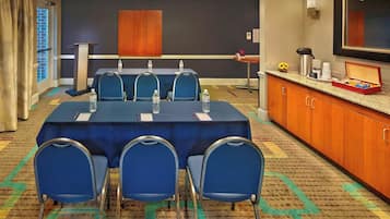 A conference room with a long table, four blue chairs, a podium, a small table with a coffee maker, and a mirror on the wall.