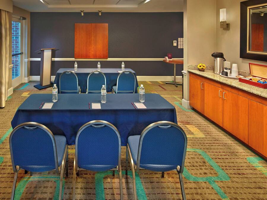 A conference room with a long table, four blue chairs, a podium, a small table with a coffee maker, and a mirror on the wall.