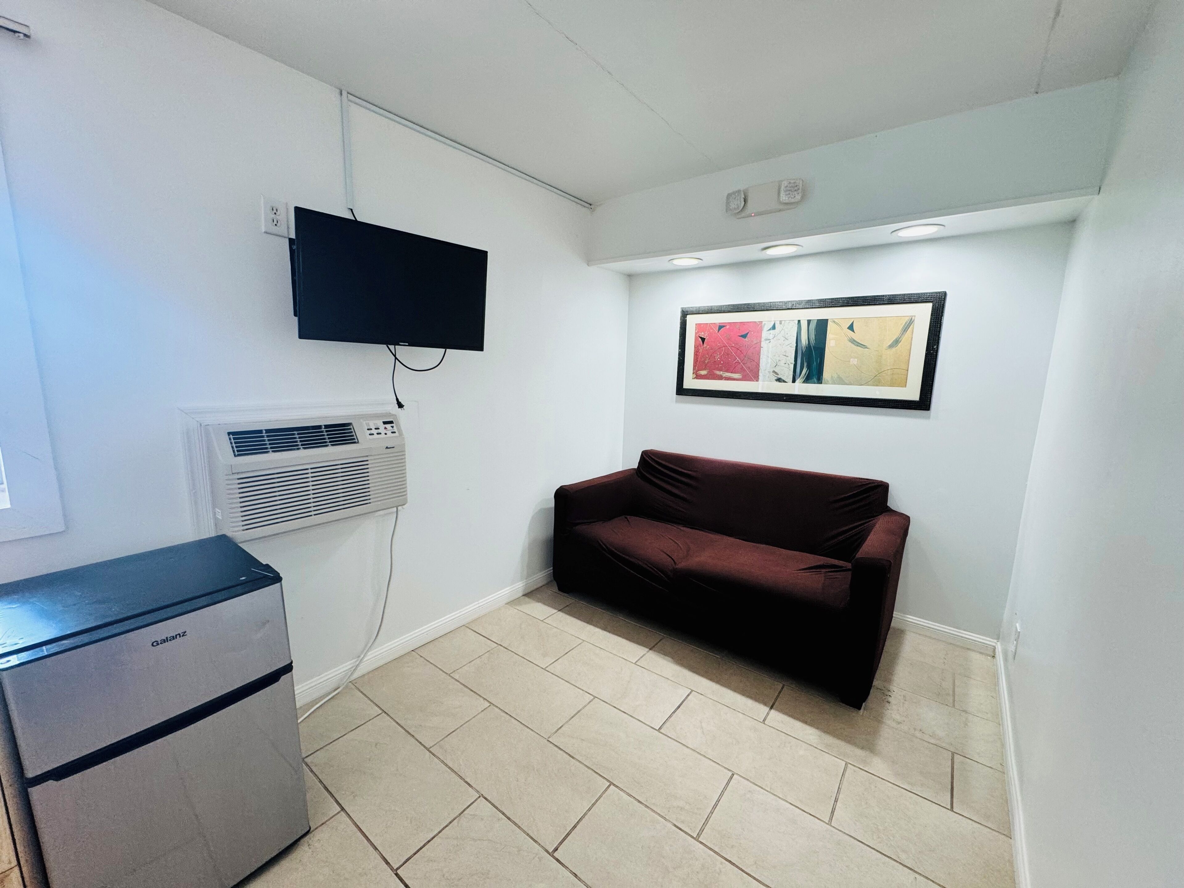 A small, clean room with a flat-screen TV mounted on the wall, a heater, a sofa, and a small refrigerator.