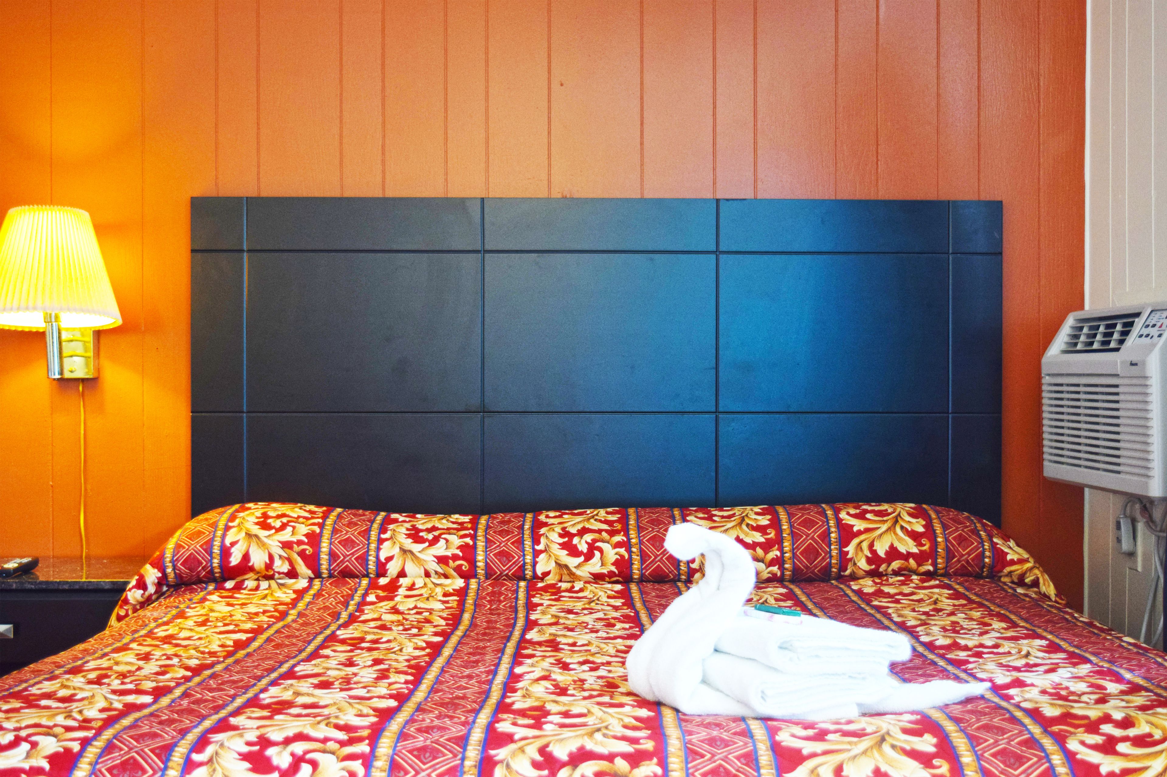A hotel room with a bed featuring a red and gold patterned bedspread, a white swan decoration, a dark headboard, a wall-mounted lamp, and an air conditioning unit.