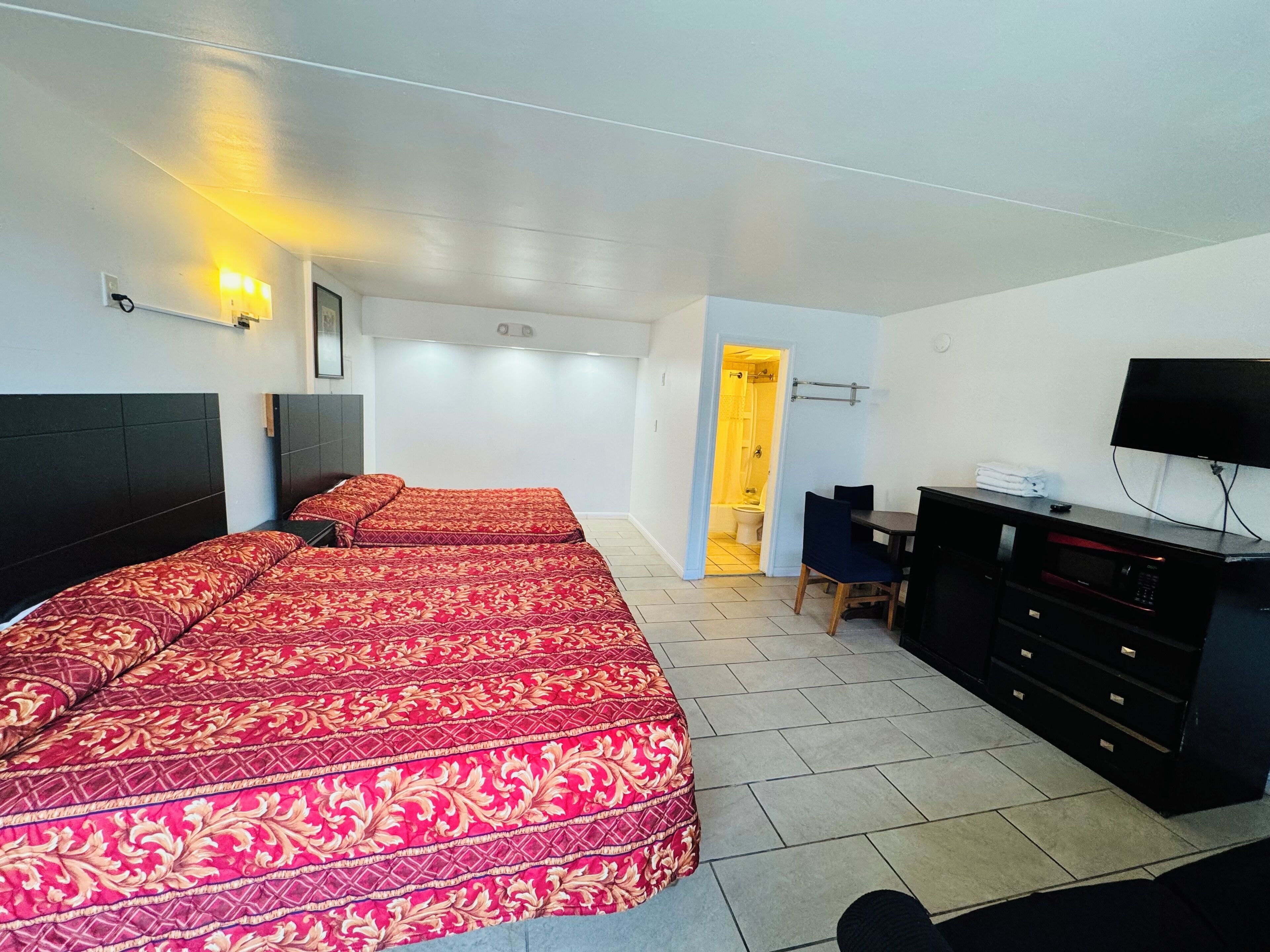Habitación de hotel con dos camas, un escritorio, un televisor y un baño visible a través de una puerta abierta.