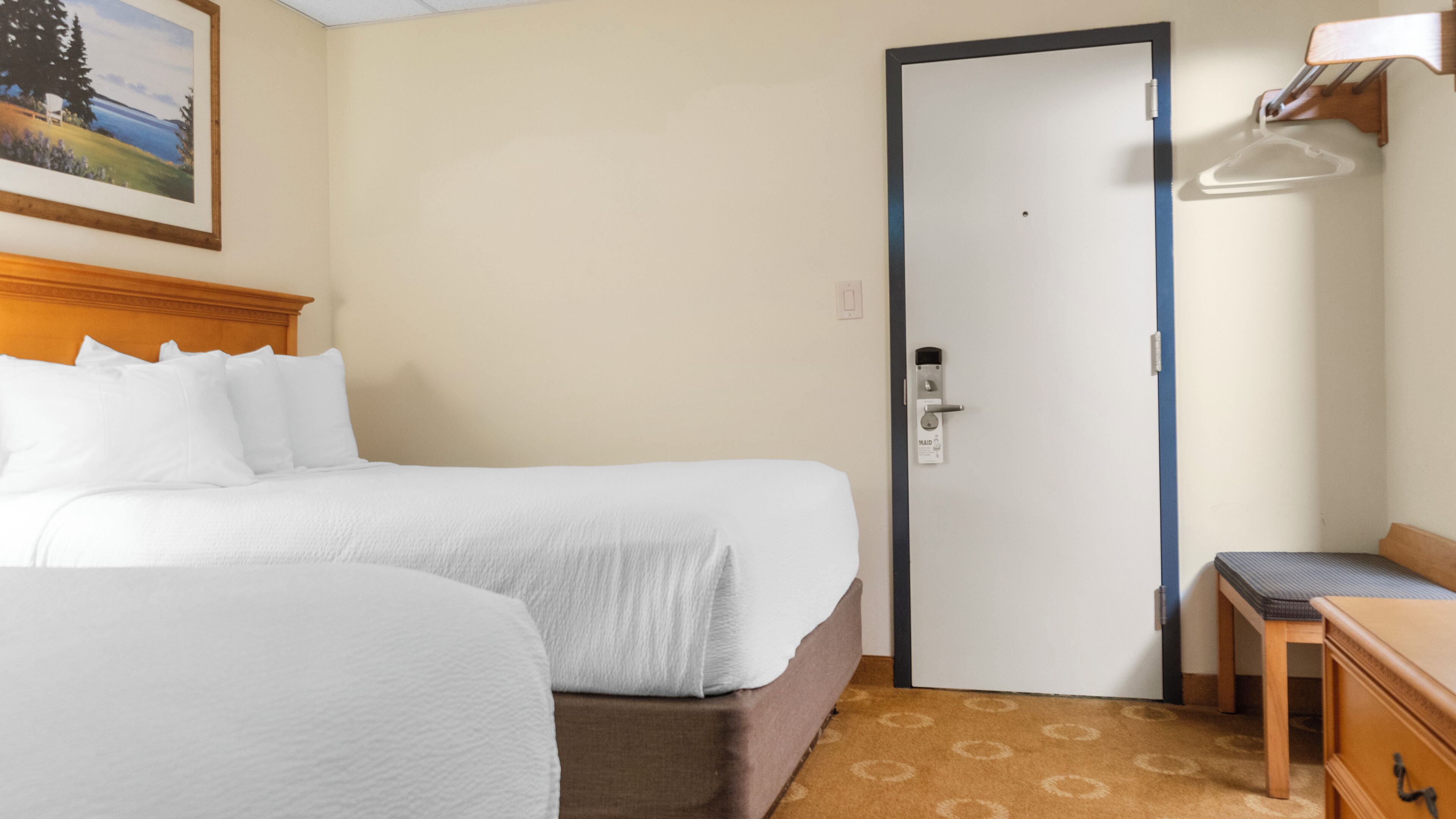 A hotel room with two beds, a wooden headboard, a painting on the wall, a door with a keycard lock, and a small table with a chair.