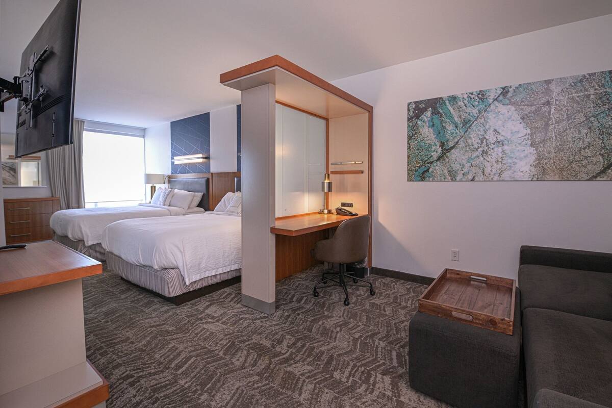 A hotel room with two beds, a desk, a chair, a TV, and a large abstract painting on the wall.