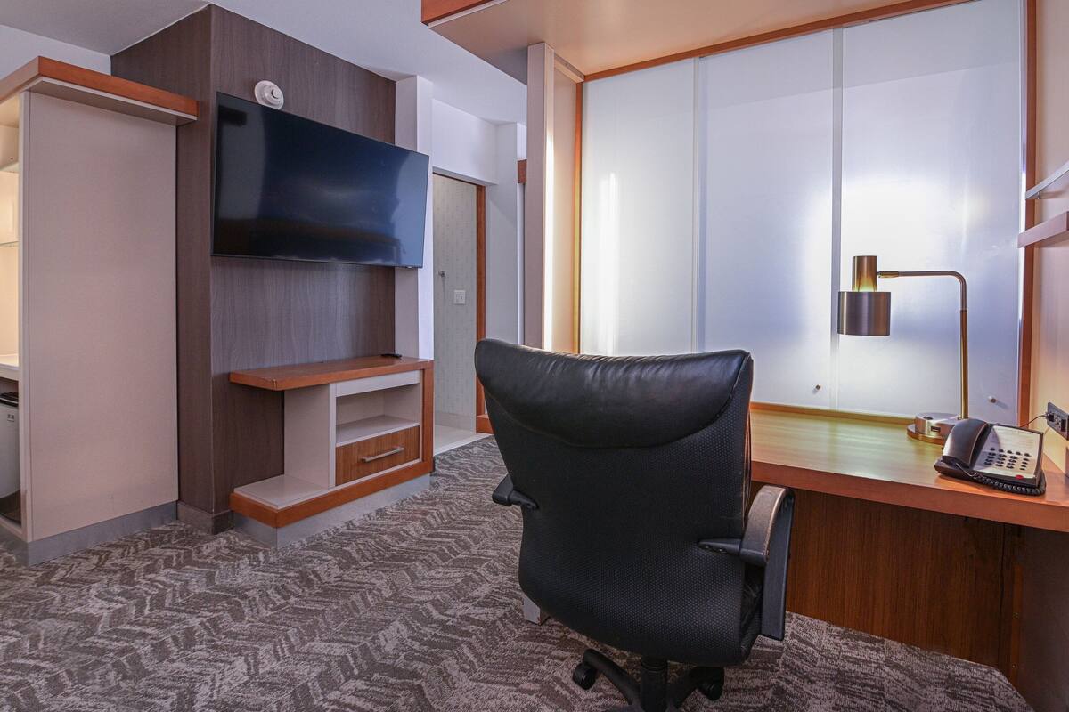 A hotel room with a desk, a chair, a flat-screen TV mounted on the wall, a telephone, and a sliding door.