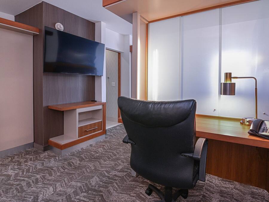 A hotel room with a desk, a chair, a flat-screen TV mounted on the wall, a telephone, and a sliding door.