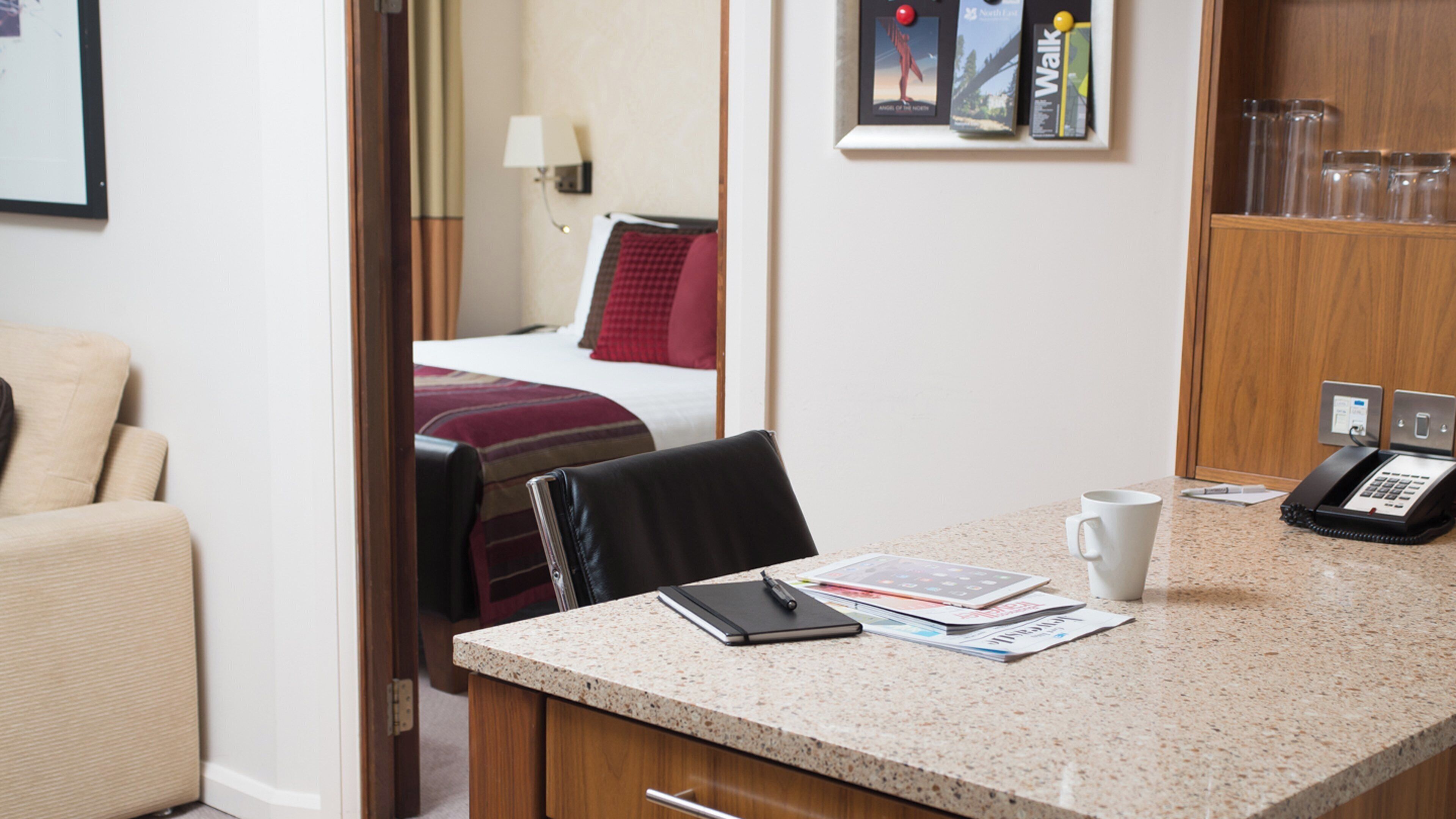 A hotel room with a bed, a desk with a phone, a chair, and a small table with a cup and some papers.