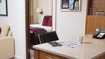 A hotel room with a bed, a desk with a phone, a chair, and a small table with a cup and some papers.