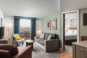 A modern hotel room with a grey sofa, a small round table, a blue armchair, a bed with white bedding, and artwork on the walls. - Staybridge Suites Newcastle by IHG (Newcastle-upon-Tyne)