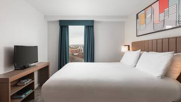 A hotel room with a large bed, a TV on a wooden stand, a city view through the window, and a painting on the wall.
