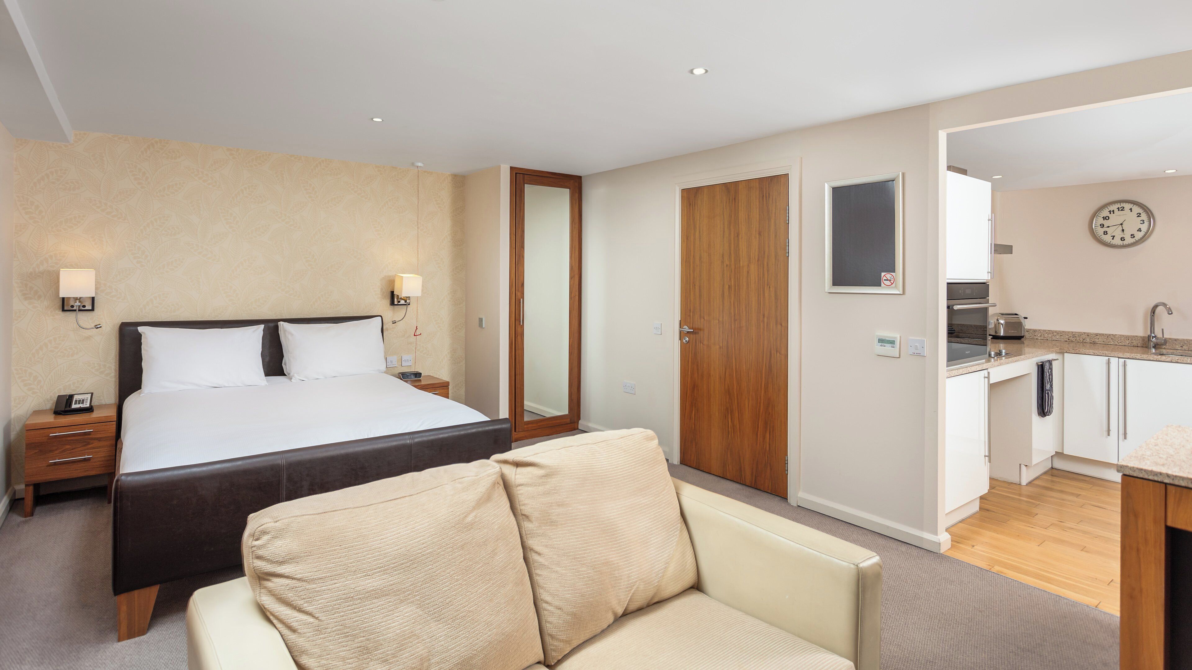 A hotel room with a bed, bedside table, lamp, and a view into the kitchen area.