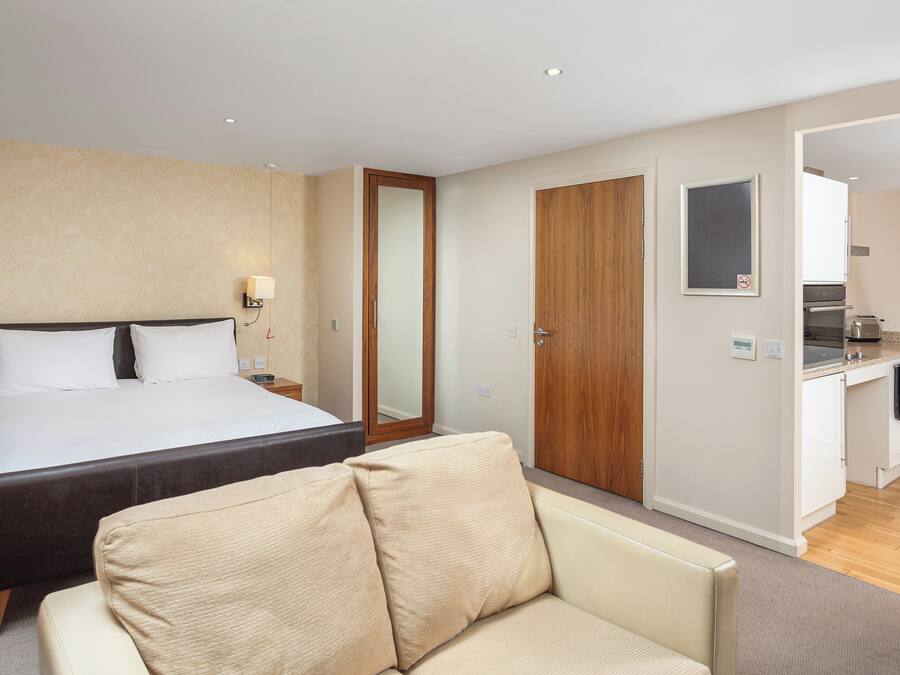 A hotel room with a bed, bedside table, lamp, and a view into the kitchen area.
