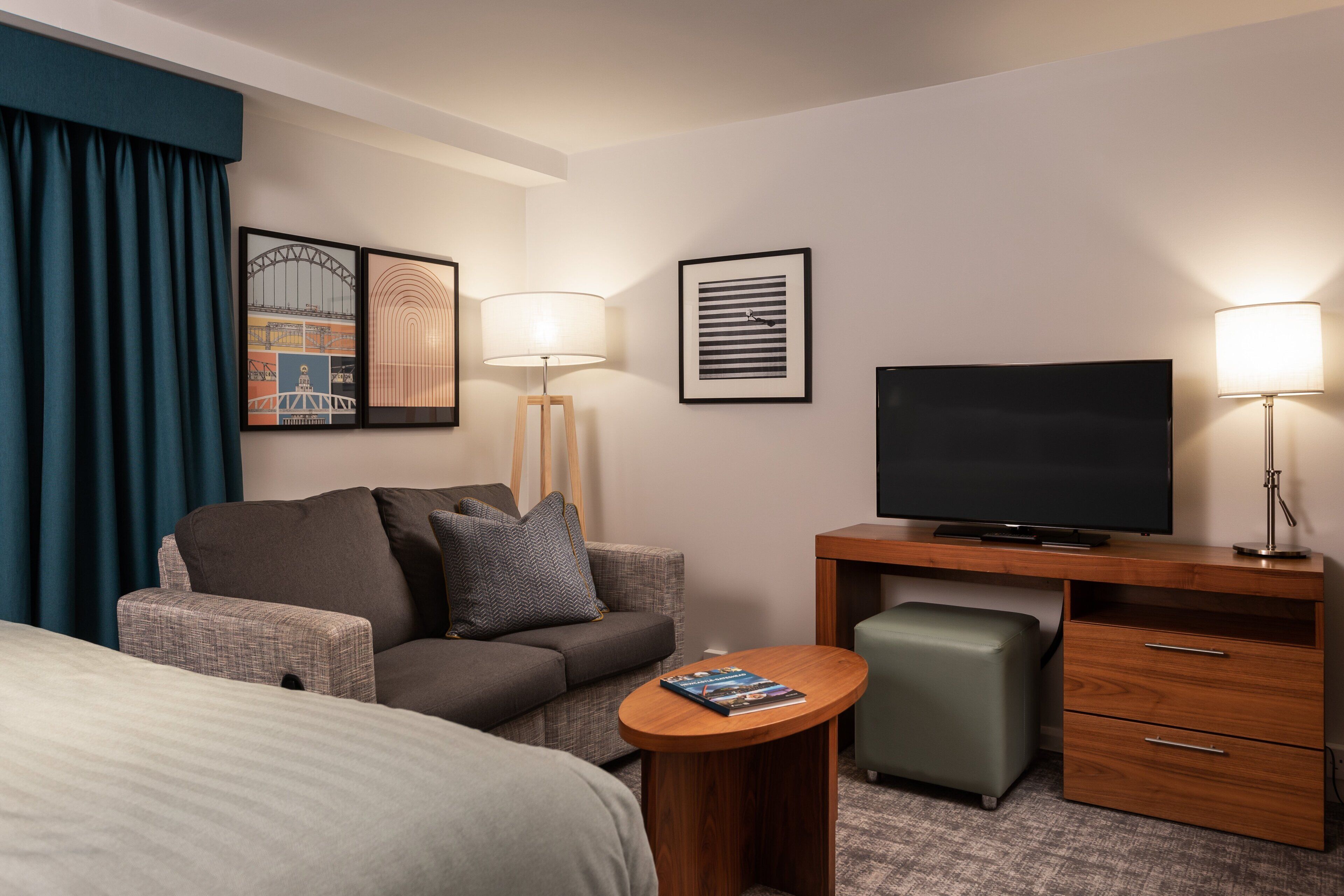 Ein Hotelzimmer mit Bett, Sofa, Schreibtisch mit Fernseher, Lampe und gerahmten Bildern an der Wand.