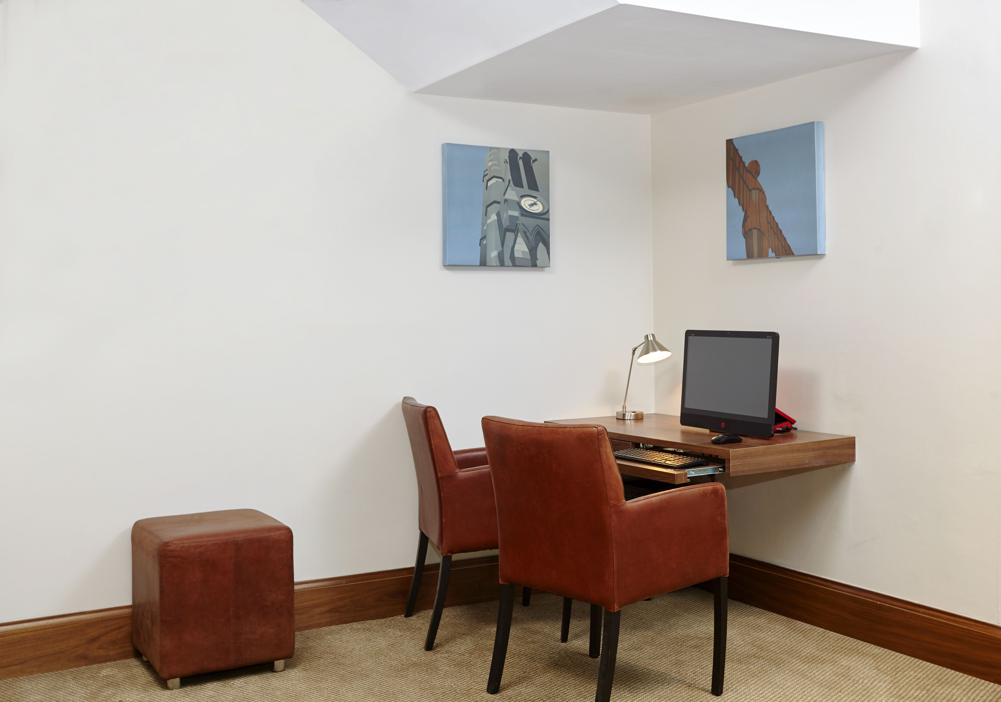 A corner office with a desk, two chairs, a computer monitor, and two paintings on the wall.