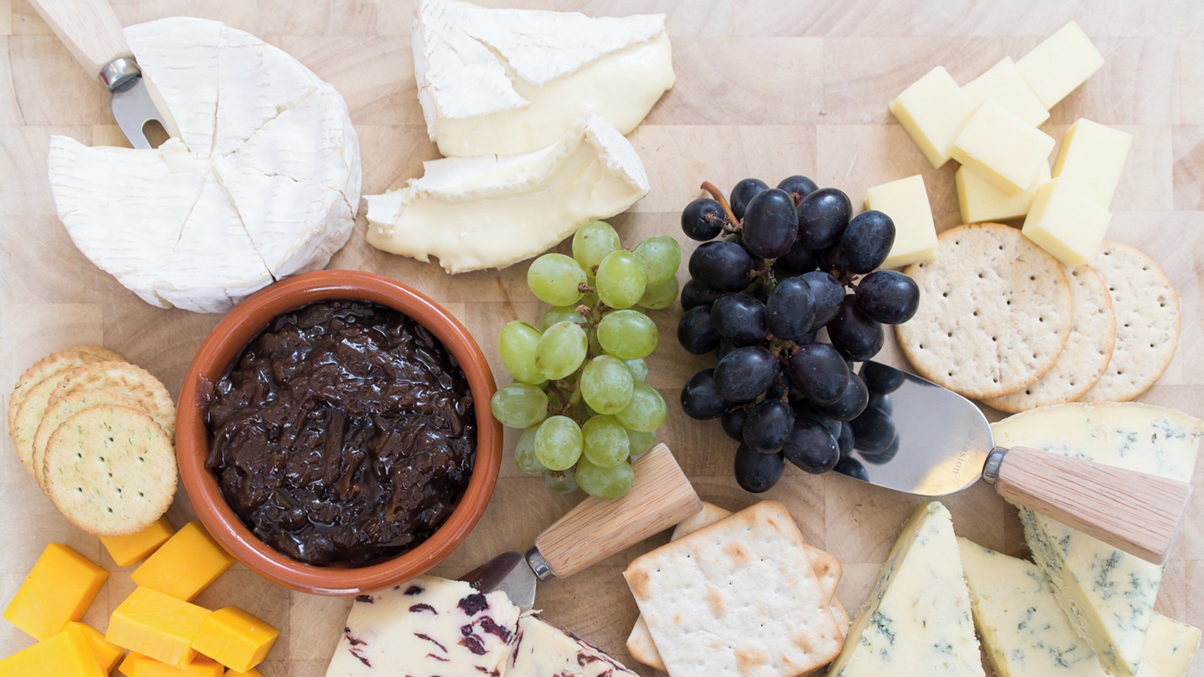 Une planche de charcuterie avec divers fromages, des raisins, des crackers et un bol de chutney.