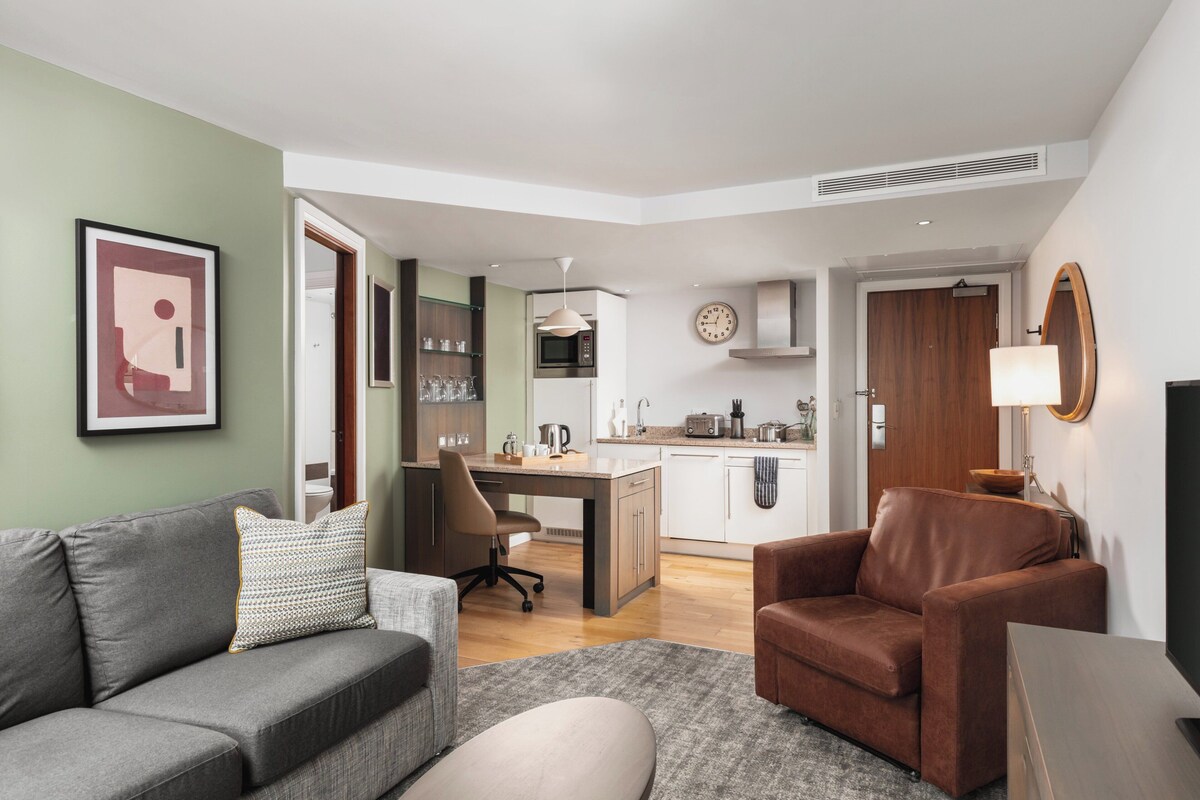 A modern living room with a grey sofa, a brown armchair, and a small table. A kitchen with white cabinets and a microwave is visible in the background.