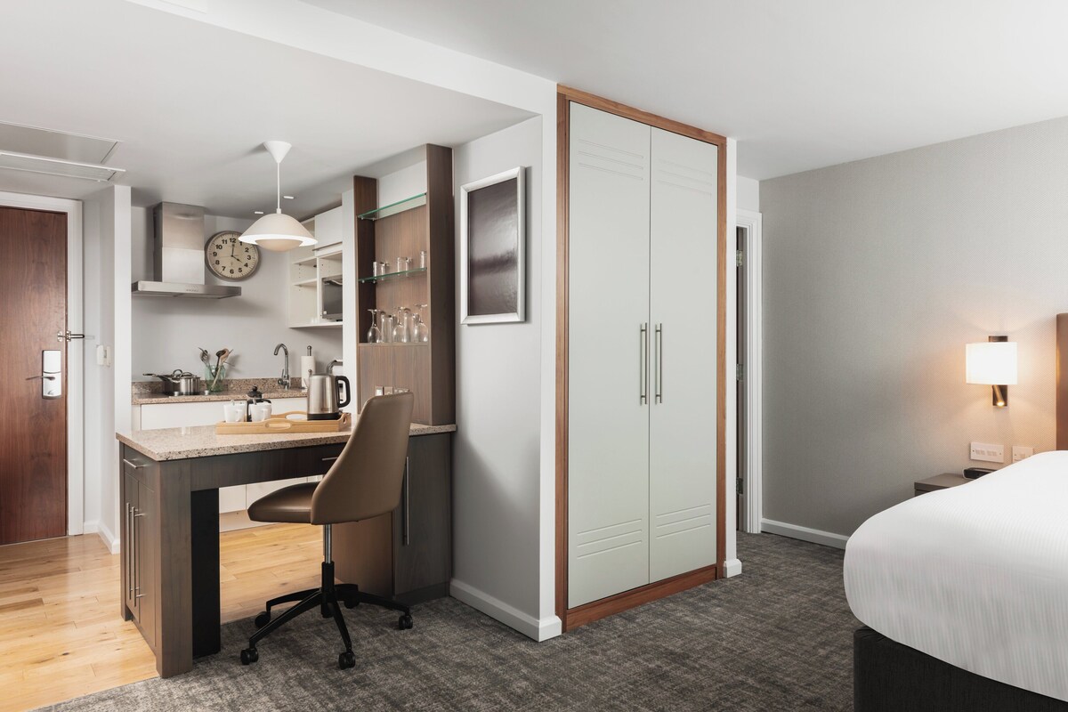 A hotel room with a kitchenette, a bed, a desk with a chair, a wardrobe, and a bedside lamp.