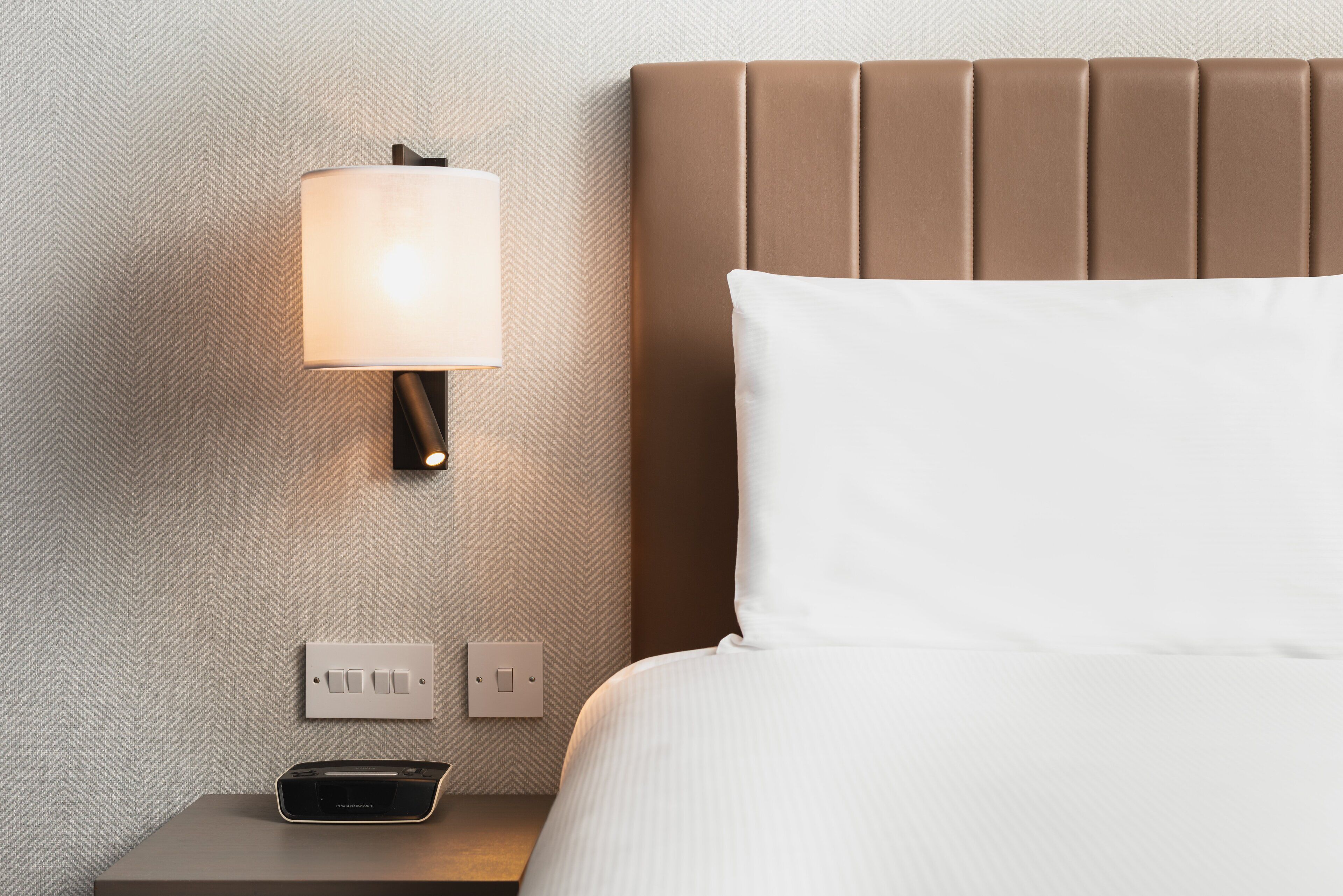 Una cama bien hecha con una almohada blanca y un cabecero marrón, una lámpara de pared y una mesita de noche con un control remoto.