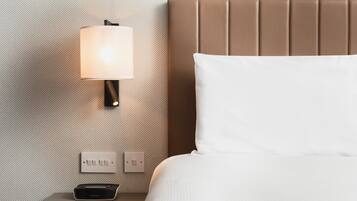Una cama bien hecha con una almohada blanca y un cabecero marrón, una lámpara de pared y una mesita de noche con un control remoto.