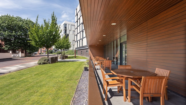 An outdoor seating area with wooden tables and chairs, a well-maintained lawn, and a modern building with large windows and a sloped roof. - Staybridge Suites Newcastle by IHG (Newcastle-upon-Tyne)