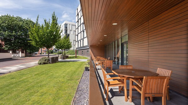 An outdoor seating area with wooden tables and chairs, a well-maintained lawn, and a modern building with large windows and a sloped roof. - Staybridge Suites Newcastle by IHG (Newcastle-upon-Tyne)