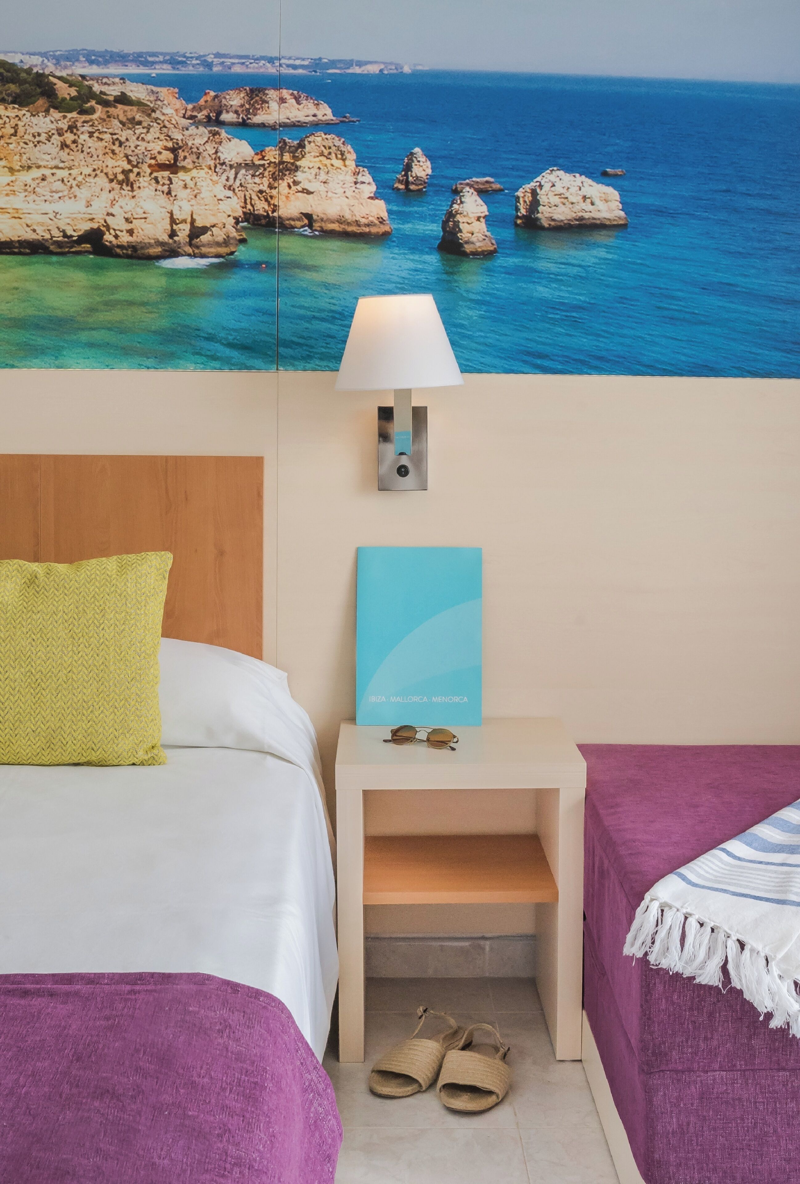 A hotel room with a bed, a nightstand with a lamp, a pair of sunglasses, and a view of the sea and rocks.