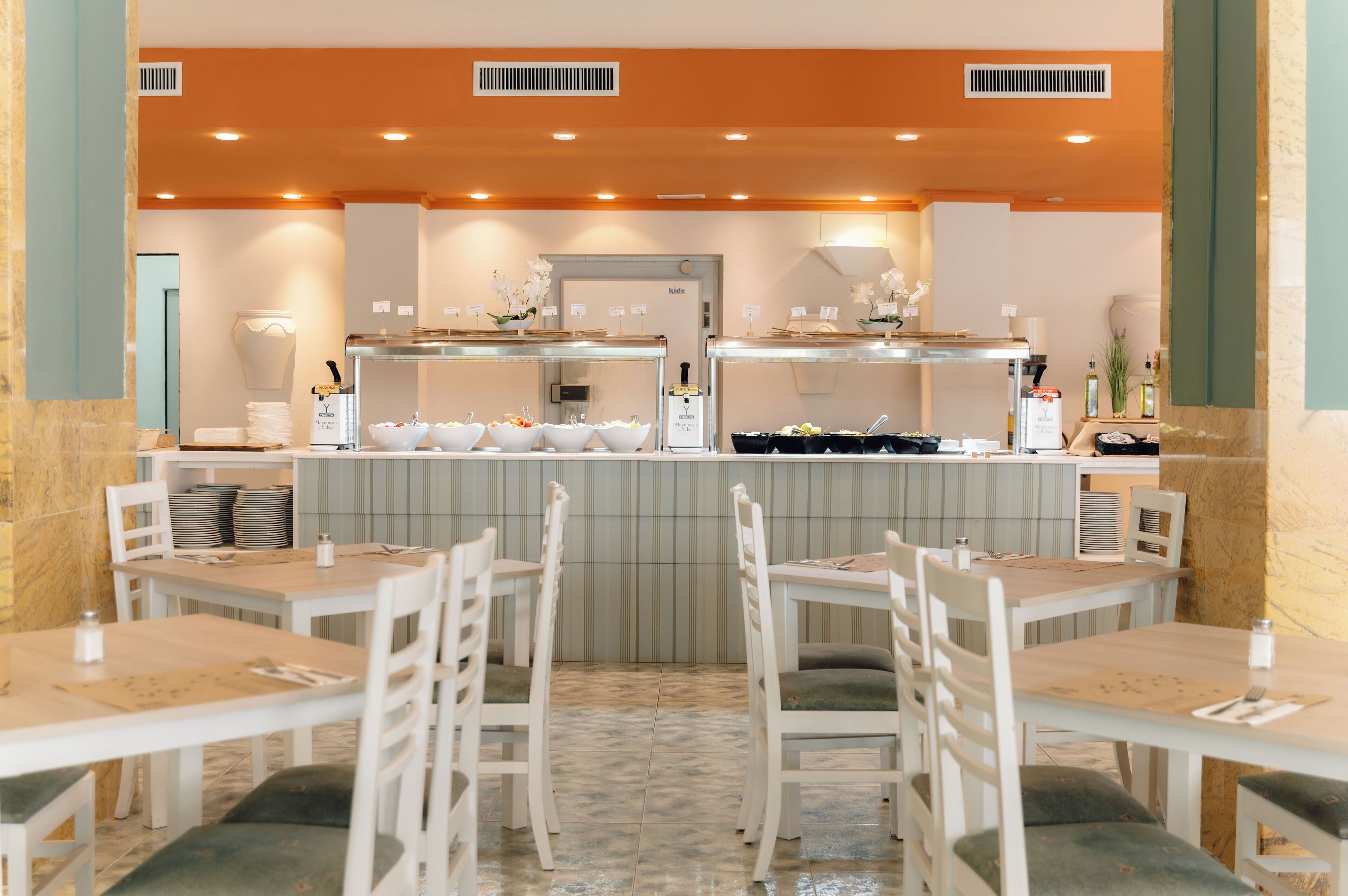 Un restaurante con mesas y sillas blancas, una barra de buffet con comida y bebidas, y un suelo de baldosas.