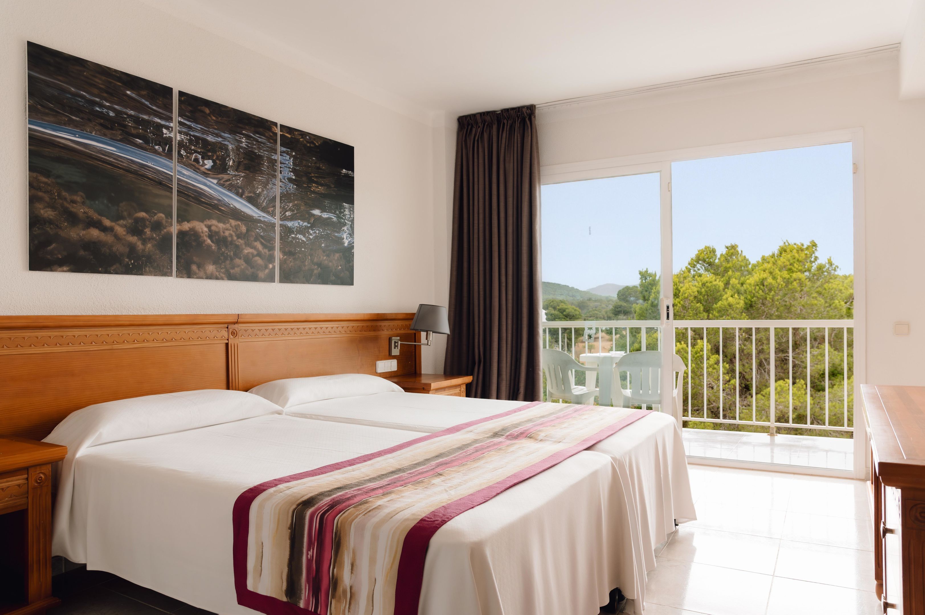 A hotel room with a large bed, a nightstand, a lamp, a view of greenery through a balcony door, and a painting on the wall.