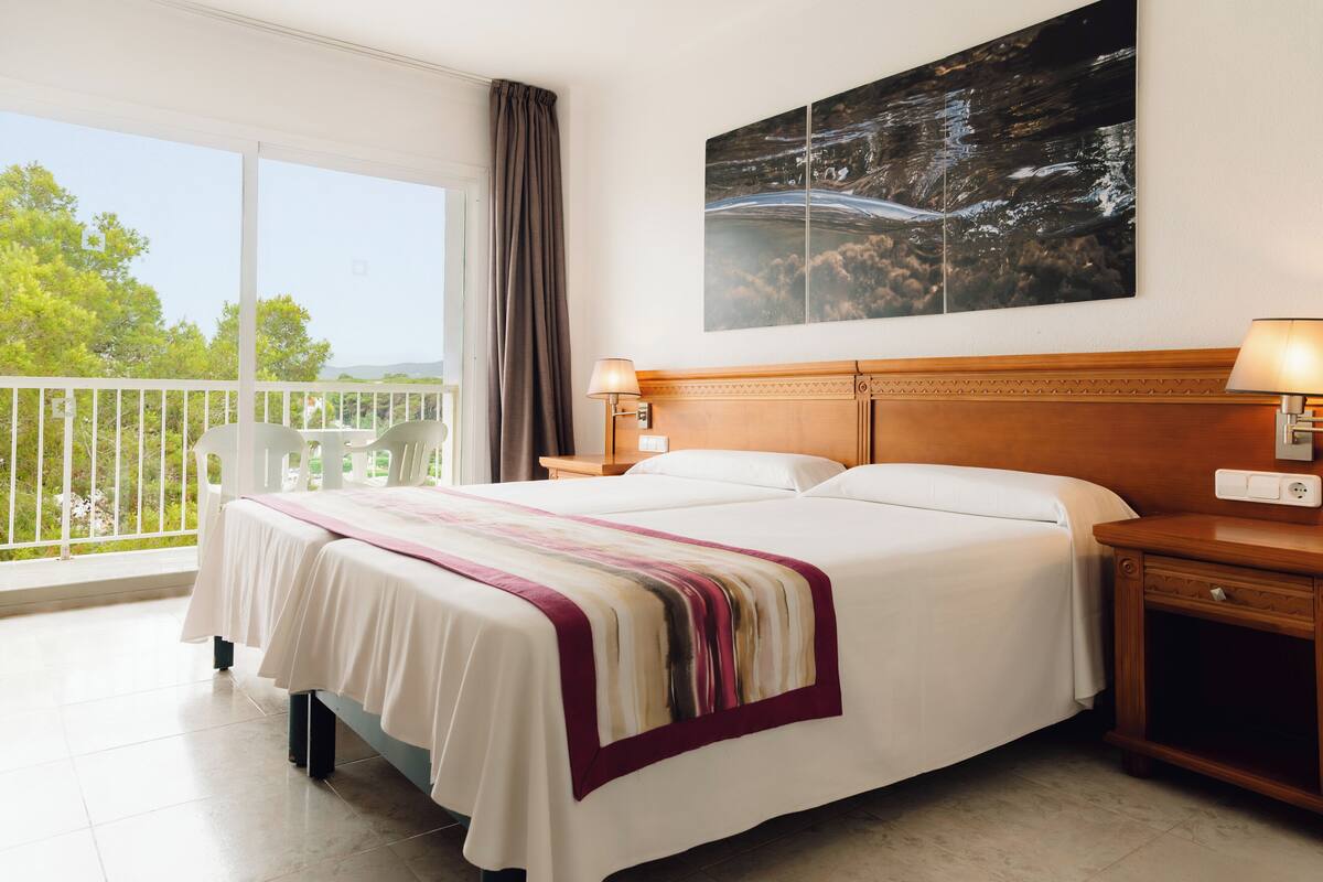 A hotel room with a large bed, a nightstand, a lamp, and a view of the outdoors.