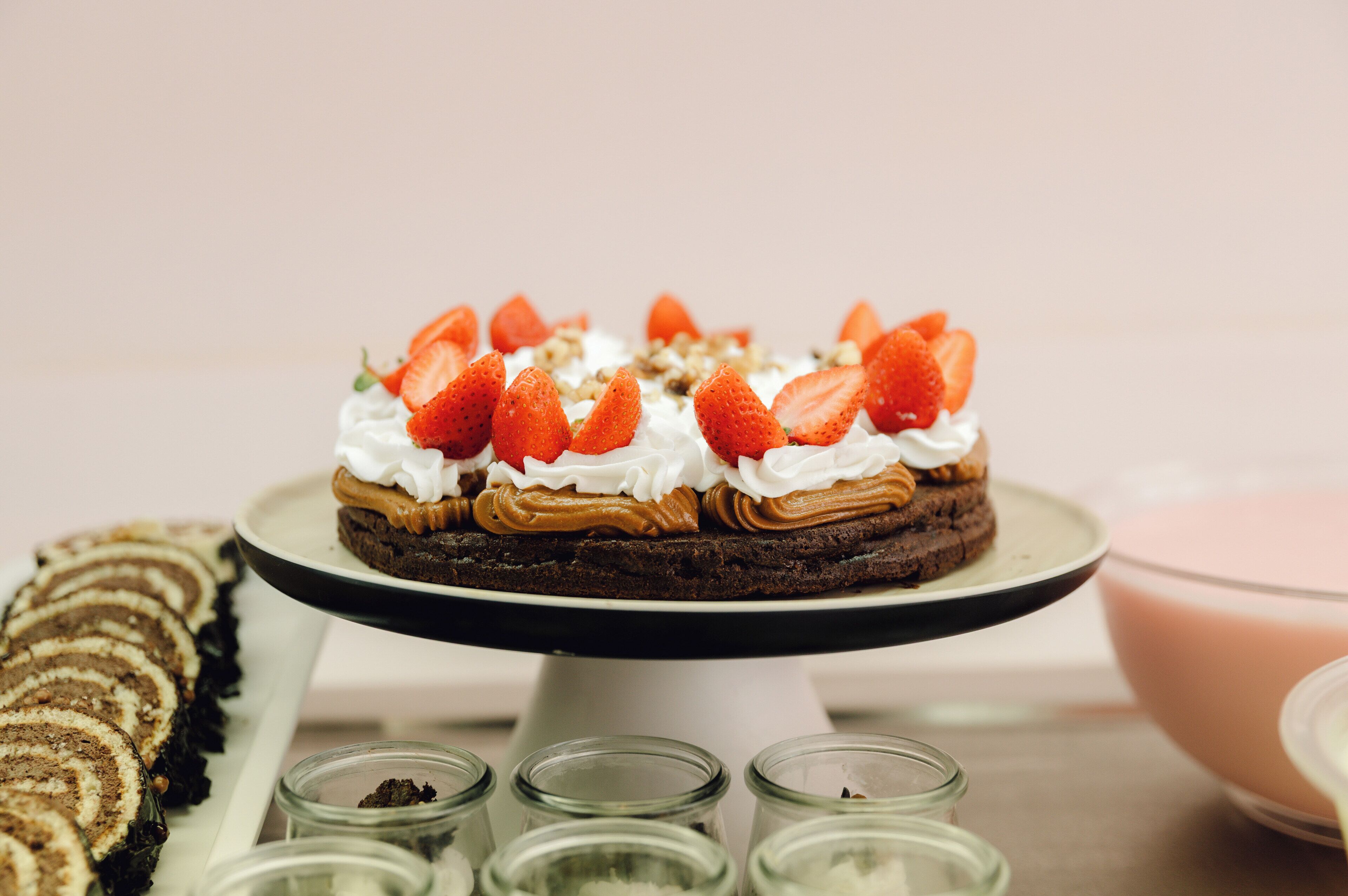 A dessert cake on a white pedestal stand with strawberries and whipped cream on top, surrounded by other desserts and small glass containers.