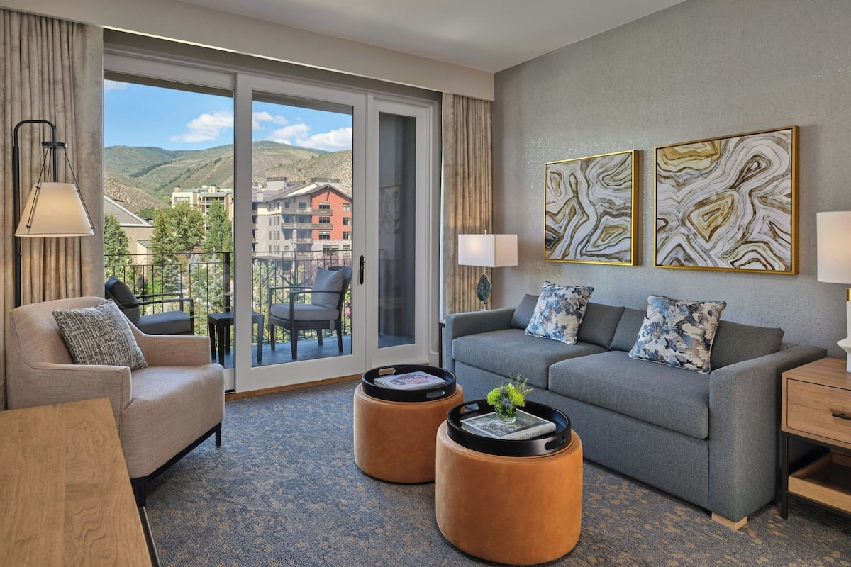 A modern hotel room with a grey sofa, two round ottomans, a wooden side table, and a balcony with a view of the mountains.