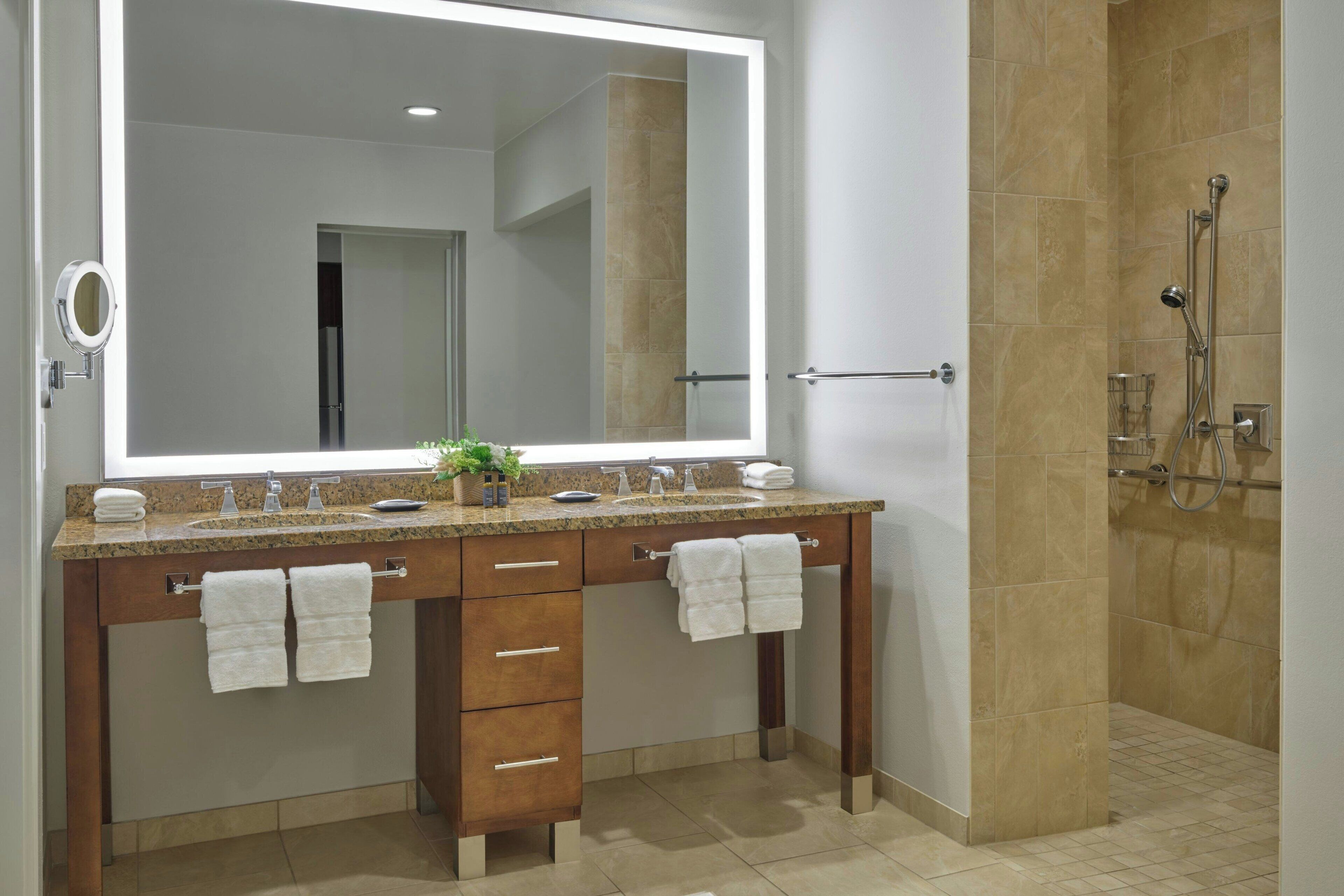 A modern bathroom with a large mirror, two sinks, a shower, and a vanity with drawers and towel racks.