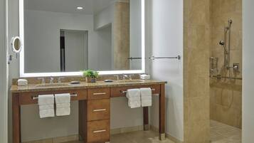 A modern bathroom with a large mirror, two sinks, a shower, and a vanity with drawers and towel racks.