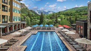 Une piscine entourée de bâtiments et dominée par un paysage montagneux, avec des chaises longues et des parasols à proximité.