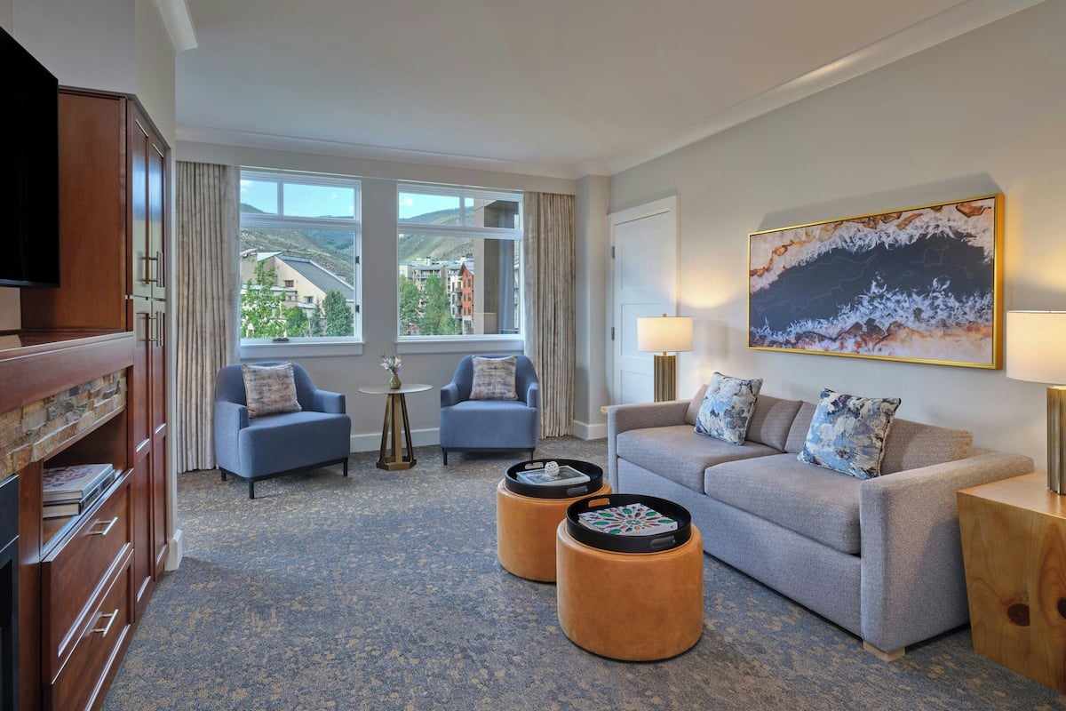 A hotel room with a sofa, two armchairs, a TV, a bookshelf, a painting, and a view of buildings and hills.