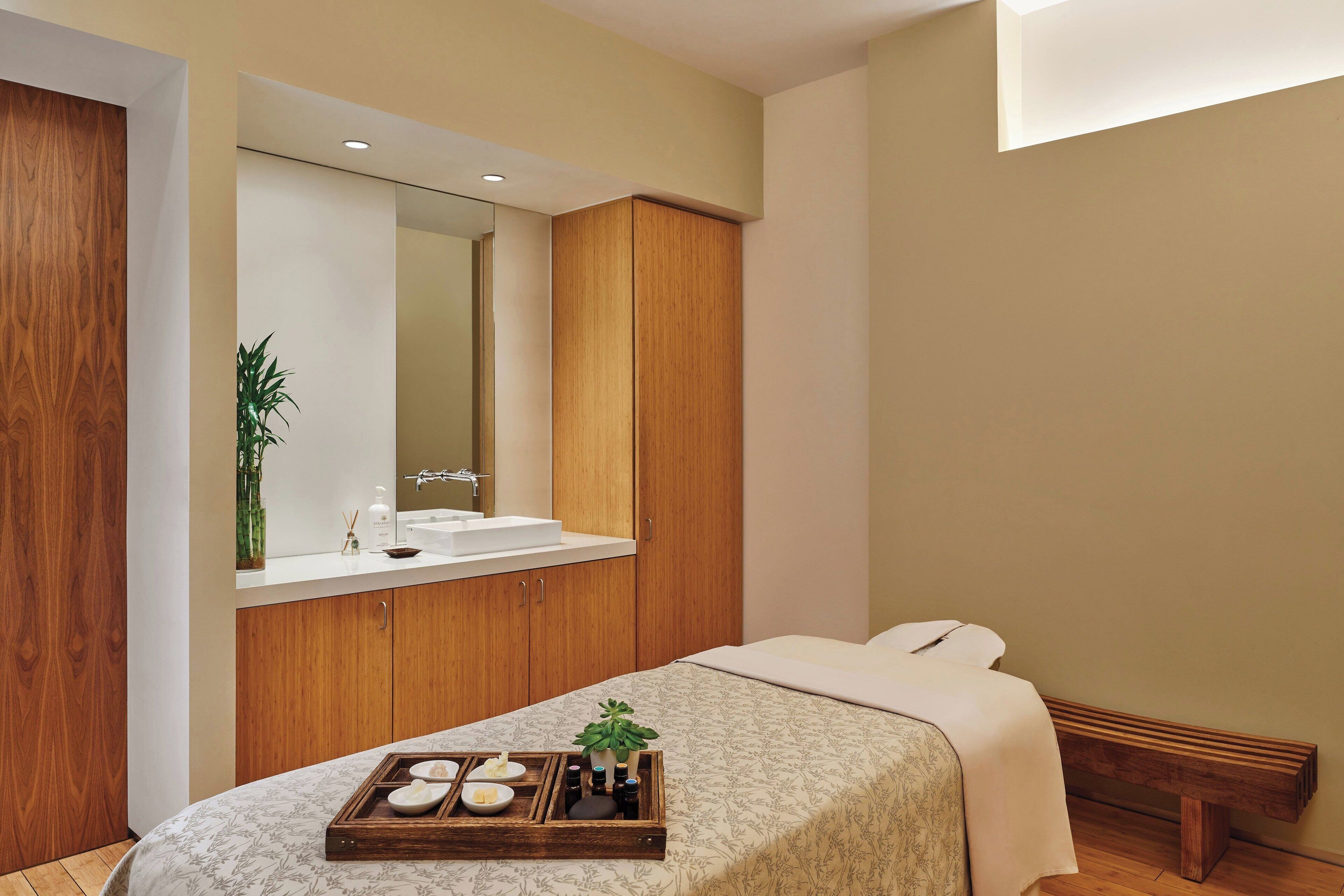 A modern bedroom with a wooden wardrobe, a bedside table with a tray of toiletries, and a large mirror above the sink.