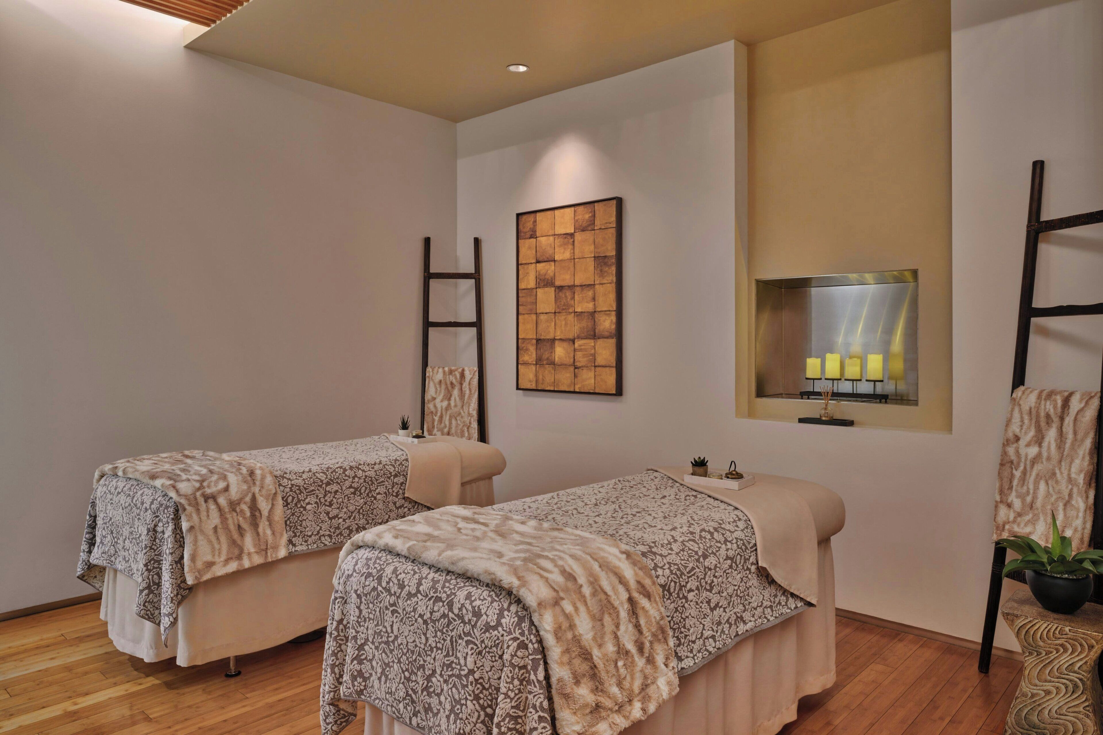 Une salle de spa équipée de deux tables de massage, d’un plancher en bois, d’une étagère murale avec des bougies et d’une plante en pot.