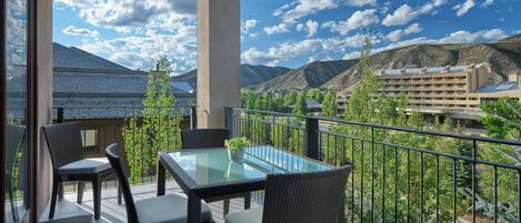 A balcony with a glass table and chairs, overlooking a mountainous landscape and other buildings.