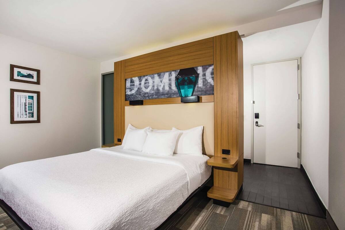 Une chambre d’hôtel avec un grand lit, une tête de lit en bois, un tableau encadré au mur et une porte sur le côté droit.