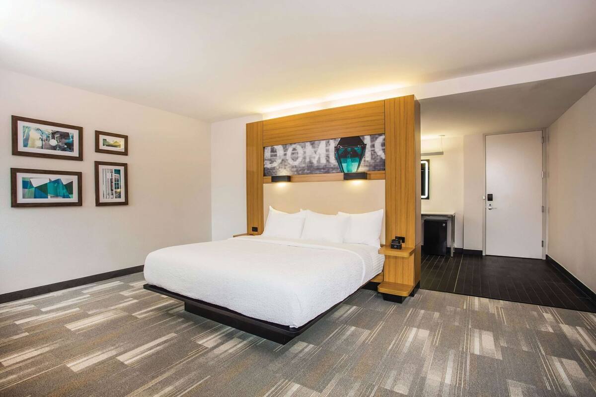 Une chambre d’hôtel avec un lit, une tête de lit en bois, un téléviseur fixé au mur et des tableaux encadrés.