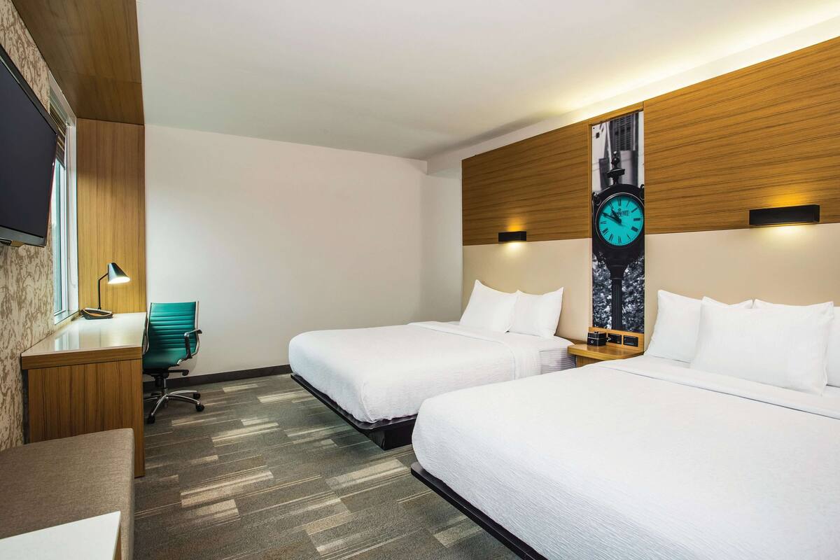 Une chambre d’hôtel avec deux lits, un bureau, une chaise, une horloge au mur et un téléviseur.