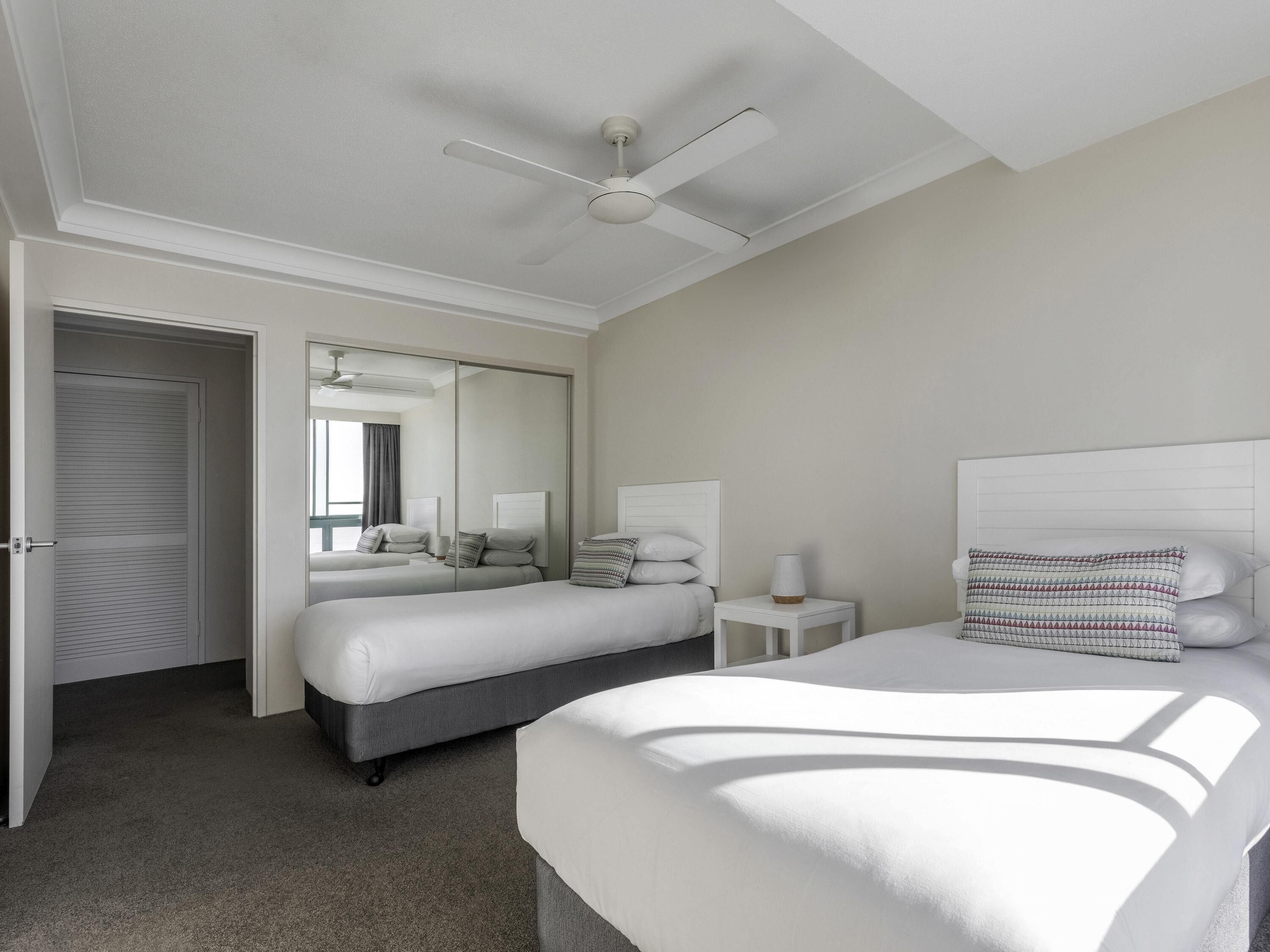 A hotel room with two beds, a ceiling fan, a mirror, and a small bedside table.