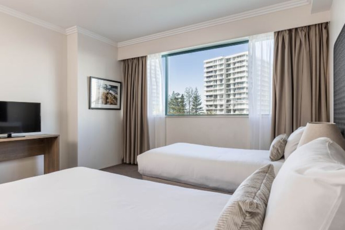 A hotel room with two beds, a TV, a window with curtains, and a framed picture on the wall.