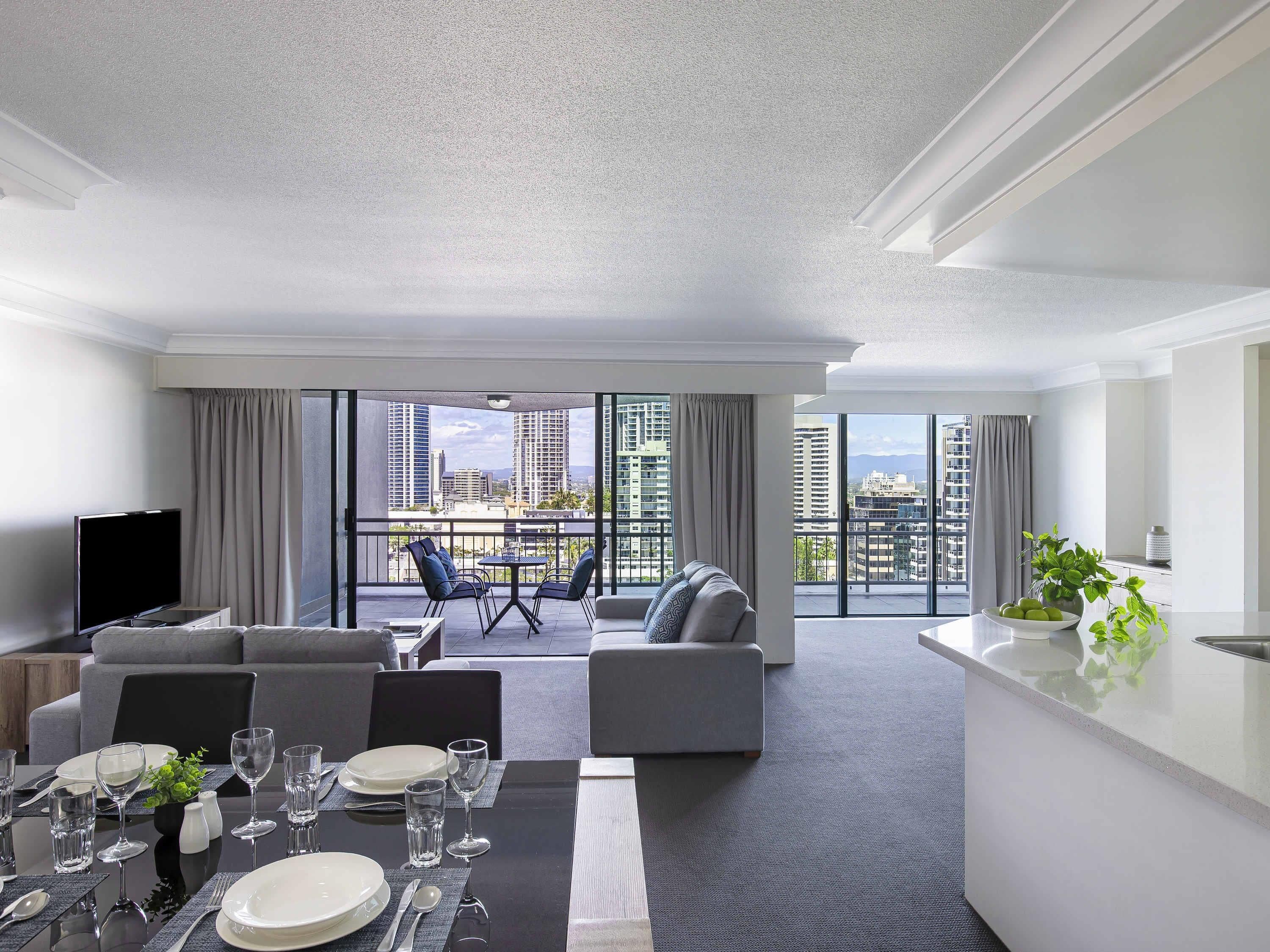 A modern hotel room with a dining area, a sofa, a TV, and a balcony with a city view.