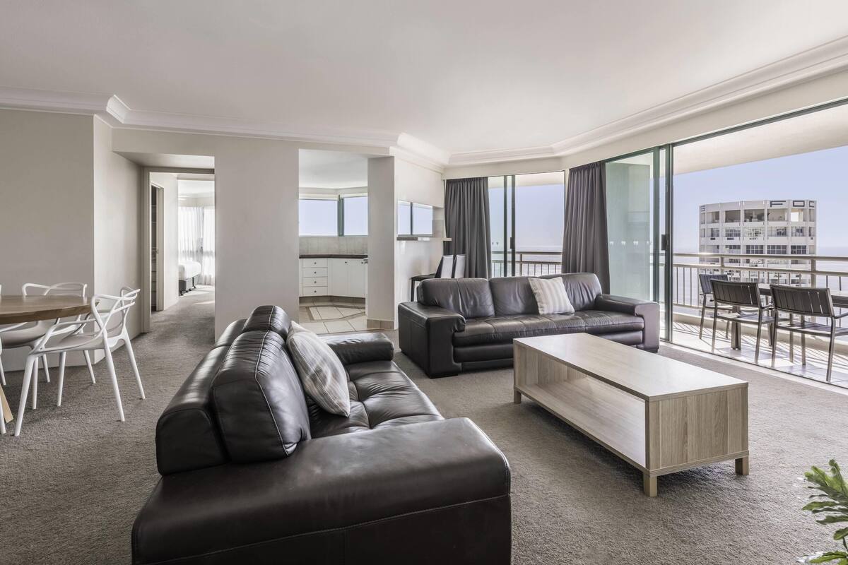A modern hotel room with a living area, a balcony with a view, and a kitchen in the background.