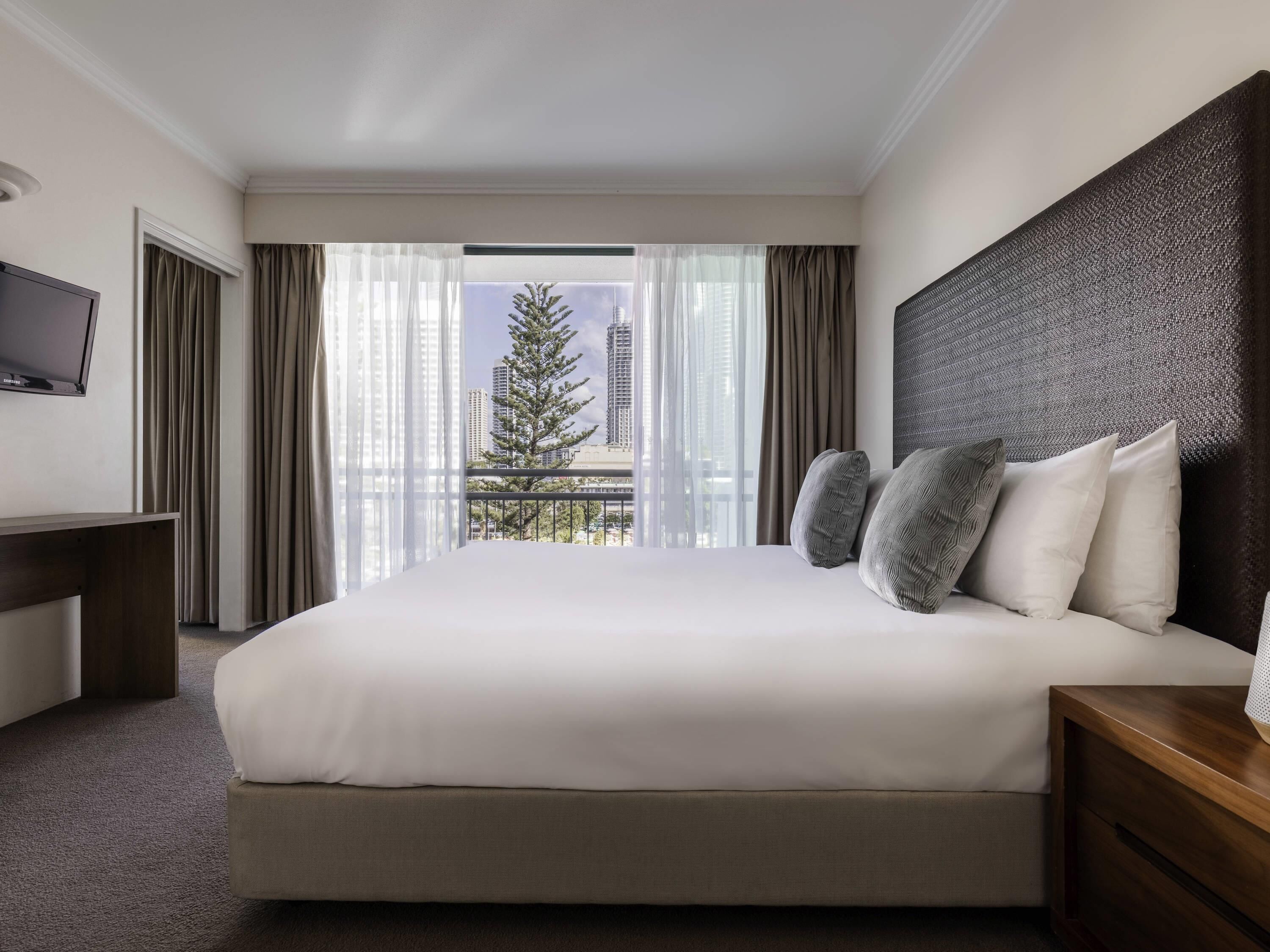 Habitación de hotel con una cama grande, un televisor a la izquierda y vistas a edificios y un árbol desde la ventana.