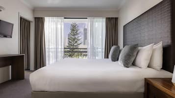 Habitación de hotel con una cama grande, un televisor a la izquierda y vistas a edificios y un árbol desde la ventana.