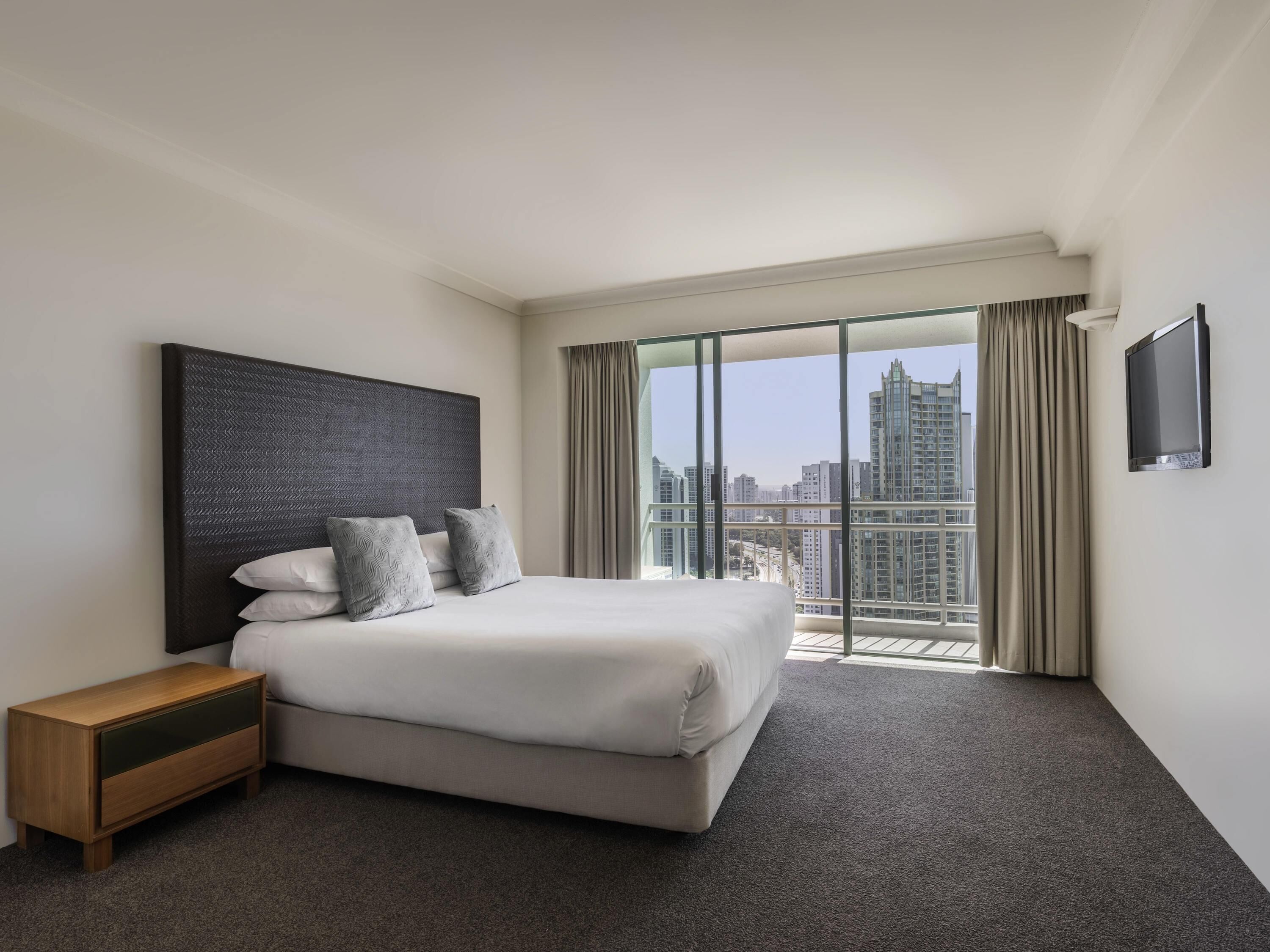 A hotel room with a large bed, a nightstand, a flat-screen TV, and a city view.