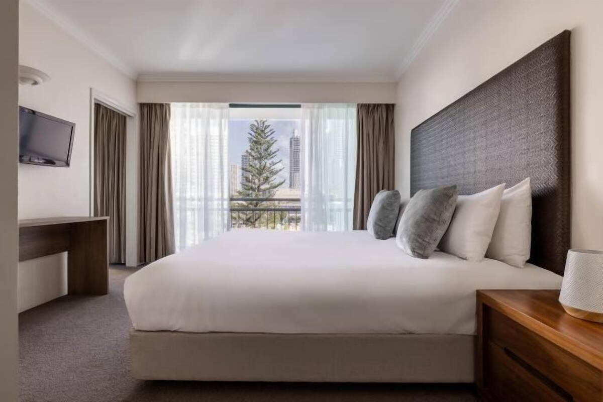 Una camera d'albergo con un letto, una televisione, un comodino in legno e vista su un albero e degli edifici.