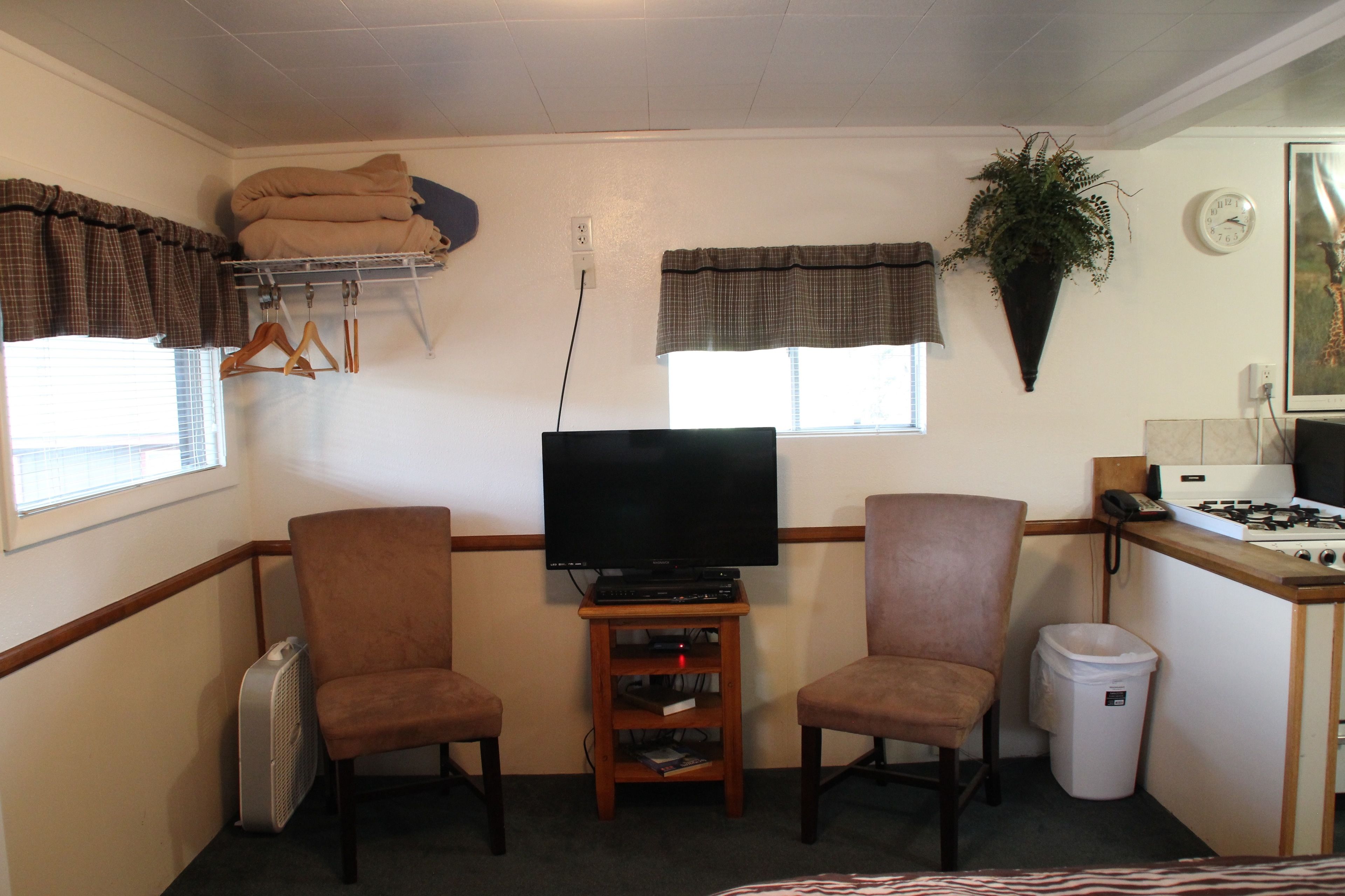 A compact living space with a television, two chairs, a small shelf, a heater, a trash bin, a stove, and a potted plant.